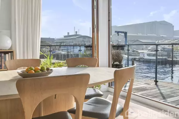 Et træspisebord med stole står ved et stort vindue, der åbner ud til en marina med både og bygninger i baggrunden. En skål med frugt er på bordet.