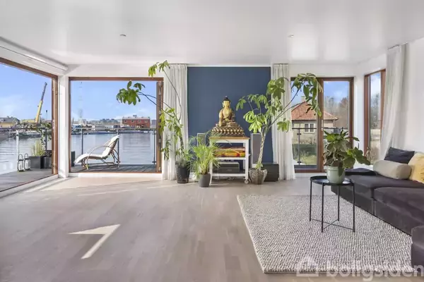 En stue med en stor Buddha-statue i centrum, omgivet af grønne planter. Sofa og tæppe er til højre, med store vinduer der viser en havneudsigt udenfor.