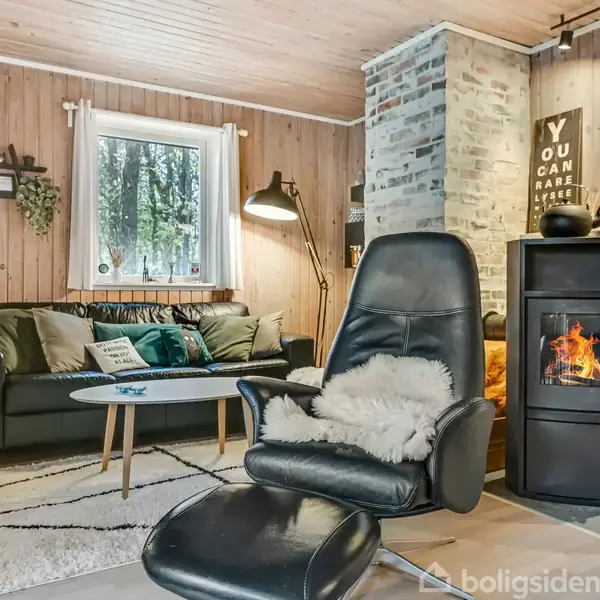 En lænestol med fåreskind står foran en brændeovn i et hyggeligt træpaneleret rum. En sort sofa med puder og et lille bord er ved siden af. Tekst på væggen: "YOU CAN HAVE IT ALL."