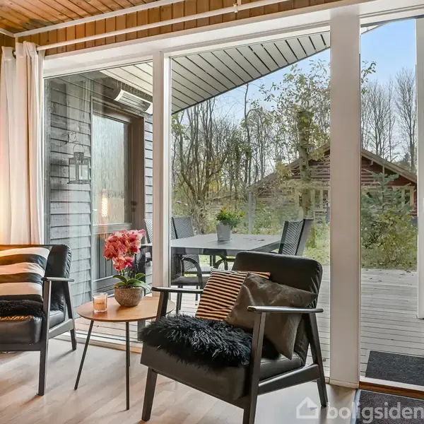 To lænestole med puder står ved et lille bord med blomster. En åben dør fører til en terrasse med spisestole og et haveområde omgivet af træer i baggrunden.