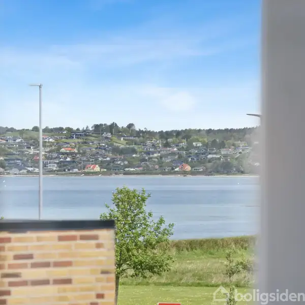 Udsigt over en rolig sø med grønne bakker og spredte huse i baggrunden. Foran ses en murstensbygning og en græsplæne. Blå himmel ovenfor.