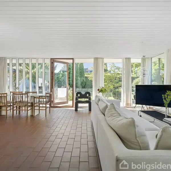 En stue domineret af en hvid sofa står overfor et TV på et sofabord. Stol og spisebord er i baggrunden. Store vinduer og glasdøre lukker lys og udsigt ind.