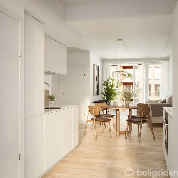 Køkken indrettet med hvide skabe og en vask, står i en lys stue-spisestue. Runde træbord omgivet af stole, pyntet med planter. Store vinduer åbner mod en balkon.