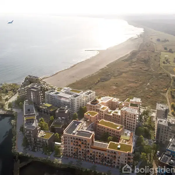 Bygninger set fra oven ligger ved kysten. Et fly svæver over havet, mens strande og grønne områder breder sig i baggrunden.