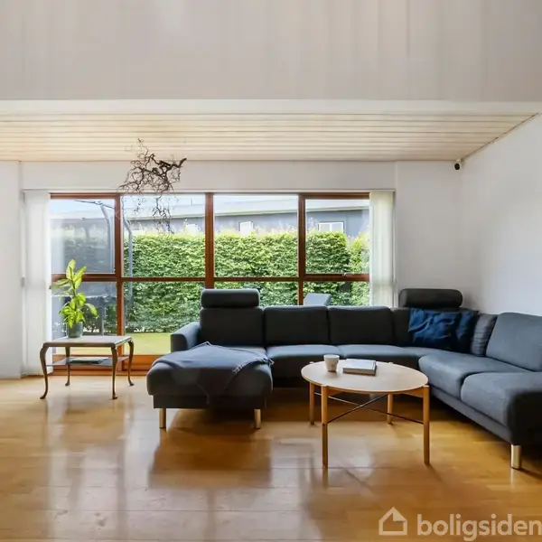 Grå sofa står ved store vinduer med udsigt til en terrasse og have. Rundt sofabord har bøger og kop. Lys træbesætning og planter i moderne stue.