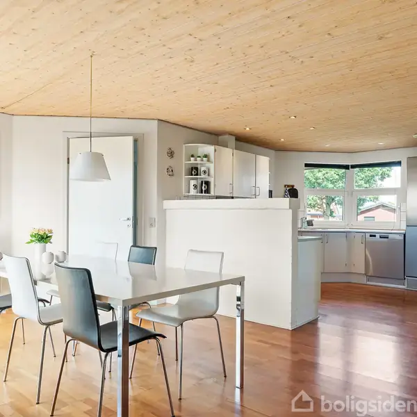 Spisebord med stole står i en moderne, lyst indrettet stue og køkken. Køkkenet har hvide skabe og stort vindue. Væggene er dekoreret med abstrakte malerier. Trægulve og loft.