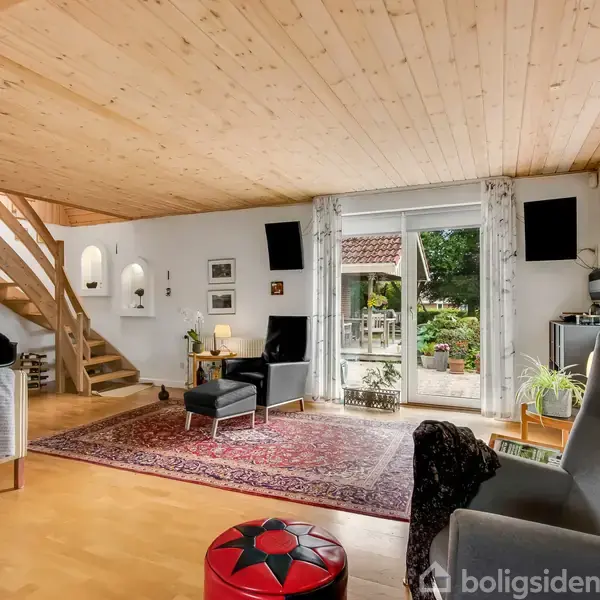 Et hyggeligt stueområde med en sort lænestol på et orientalsk tæppe, nogle planter og en stor glasdør, der åbner ud til en terrasse. Trætrapper fører til en øvre etage.