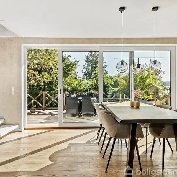 Et rektangulært spisebord med seks polstrede stole står i et lyst rum. Store glasdøre åbner op til en terrasse med grønne træer udenfor. Hængende lamper over bordet.