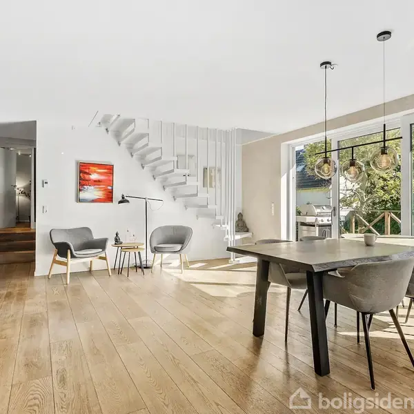 Et moderne spisestueområde med et stort træbord og seks stole, er placeret ved siden af store vinduer, der åbner ud til en terrasse med grøn udsigt. En trappe fører opad langs væggen.