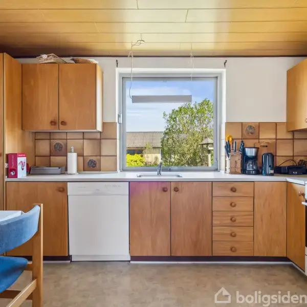 Køkken med træskabe står stille. I midten ses en vask under et vindue, omgivet af opbevaring og hvidevarer. Bord til venstre, lysindfald udefra skaber en varm atmosfære.