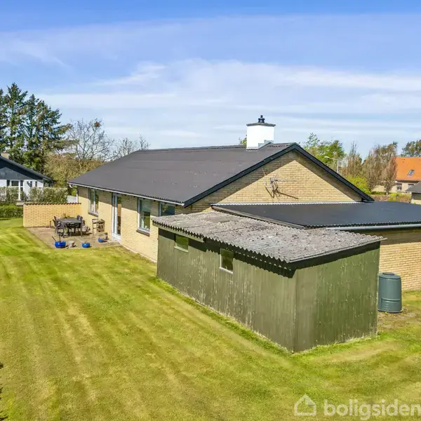 Et gult murstenshus med sort tag står stille i en grønt, velplejet have. En stor garage ses ved siden af, og der er trampolin og havemøbler i baggrunden.