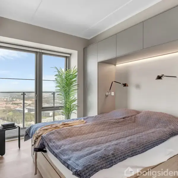 En dobbeltseng med lilla sengetøj er centralt placeret under indbyggede overskabe med læselamper. Ved siden af står en grøn plante, og en glasdør åbner mod en balkon med udsigt.