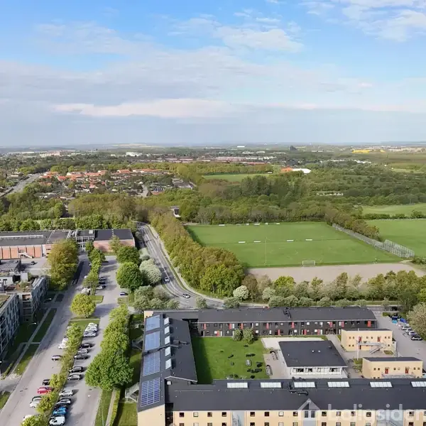 En stor park fyldt med grønne træer ligger ved siden af et boligområde med moderne bygninger. I baggrunden ses åbne marker og en blå himmel med få skyer.