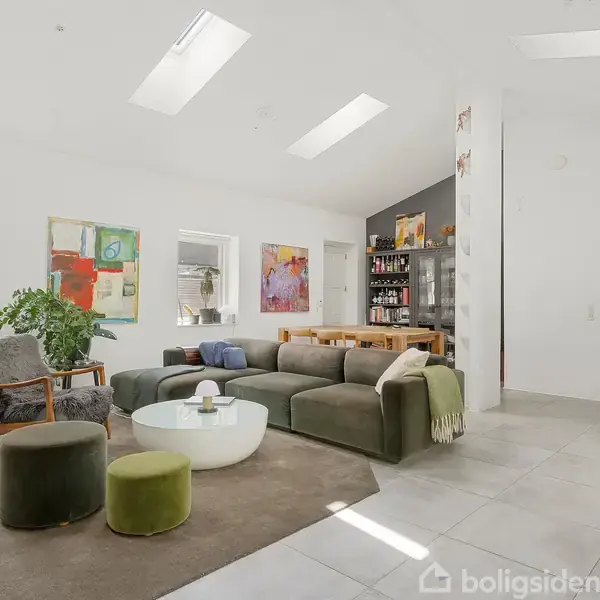 En moderne stue med grøn sofa og lænestol omkring et hvidt sofabord. Store vinduer og klinkegulv skaber lys, rummelig atmosfære. Farverige malerier hænger på væggen, og planter dekorerer rummet.