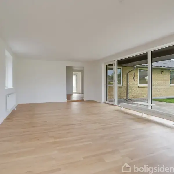 Stue med lyst trægulv og store vinduer, der giver udsigt til en terrasse og en græsplæne. Væggene er hvide, og der er en radiator under vinduet.