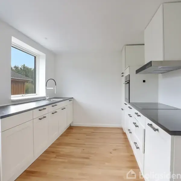 Et lyst køkken med hvide skabe langs væggene er tomt og ryddeligt. En stor, firkantet vindue til venstre giver udsigt til det grønne udenfor. Gulvet er af lyst træ.