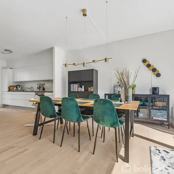 Spisebord med grønne stole står i spisestue med trægulv og moderne indretning. Køkkenet er i baggrunden. Reoler med bøger og dekorationer placeret ved væggen.
