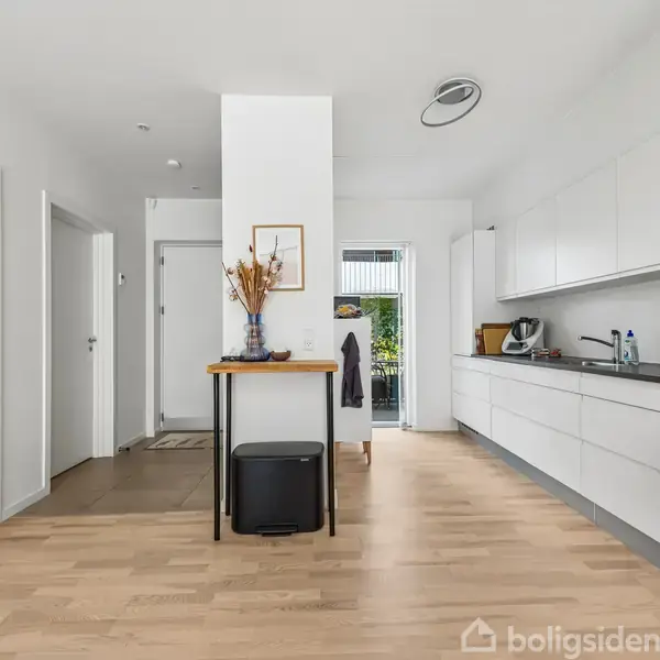 Lyst køkken med hvide skabe og indbygget ovn langs højre side. Et bord med vase og pynt står centralt. Døre fører til andre rum. Gennemsigtig dør åbner til have.