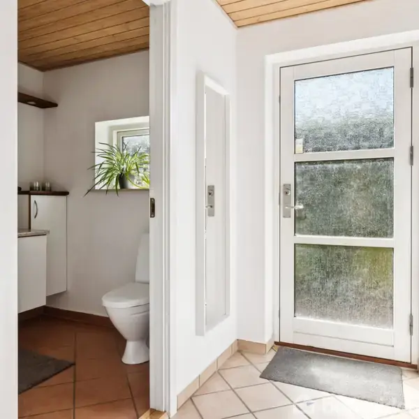 Dør til entre fører til badeværelse med toilet. Badeværelset har hvid væg, flisegulv og lille vindue med plante. Dørmåtte ligger ved glasdør med mønstret ruder indendørs.