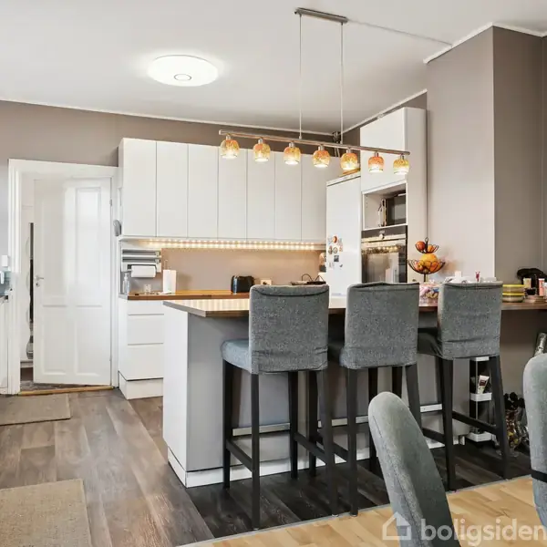 Et moderne køkken med hvide skabe, en stor køkkenø med grå barstole, og hængende lamper. Trægulve skaber en varm atmosfære, og en klokke hænger på væggen nær døren.