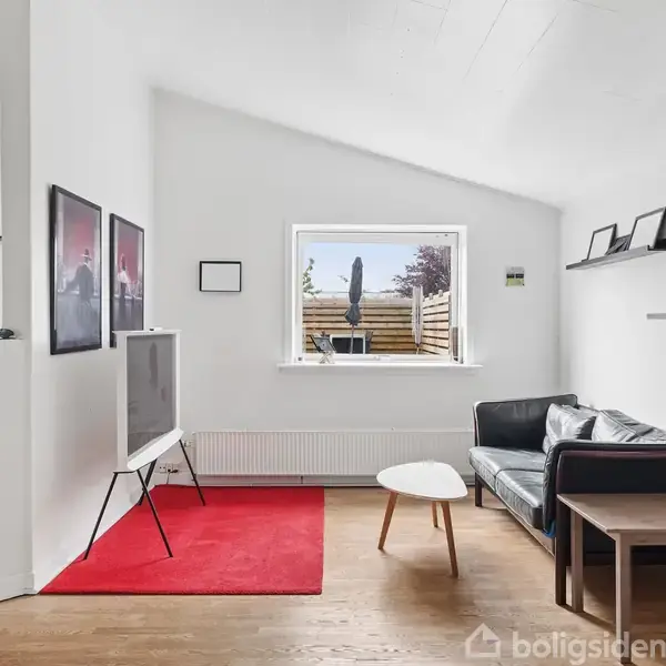 TV på stativ står ved rød måtte i stue med hvidmalede vægge. Sorte sofaer og småborde med pynt omringer rummet. Væggen har hylder og kunstværker. Vindue viser terrasseudsigten.