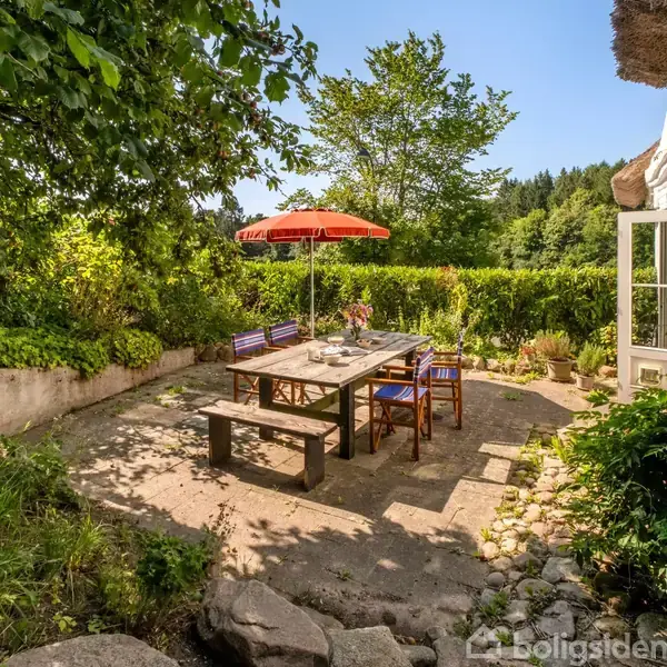 En udendørs terrasse med et stort bord og stole under en rød parasol. Terrasseområdet er omgivet af frodig vegetation og træer, der skaber en hyggelig og skyggefuld atmosfære.