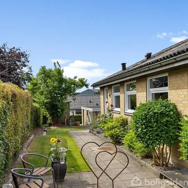 En gul murstensbungalow med store vinduer står langs en flisebelagt sti. Ved siden af stien er en have med blomster og buske. I baggrunden er en blå himmel med få skyer.