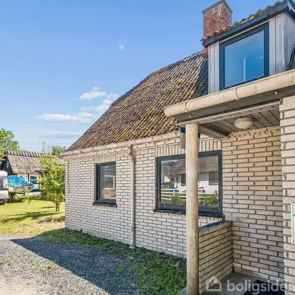 Et hus med hvide mursten står stille i en have med græsplæne. Postkassen er ved hoveddøren, og tallet "16" er synligt. En campingvogn ses i baggrunden.