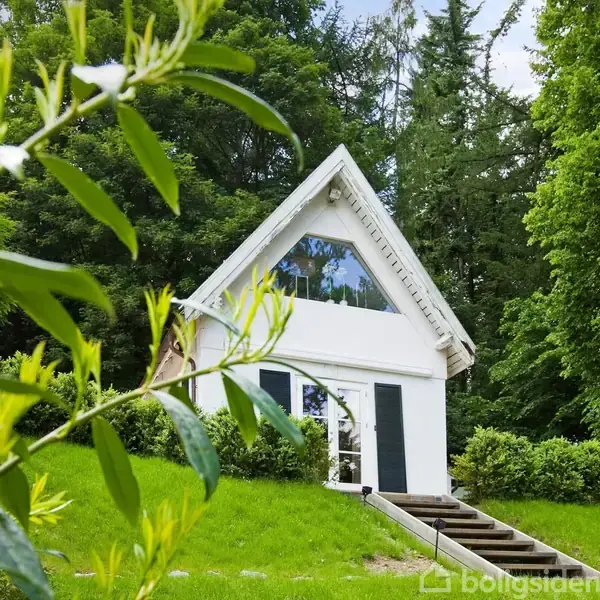 Et lille hvidt hus med trekantet tag står stille omgivet af frodige træer og buske. En græsplæne og en række trin leder op til husets indgang.