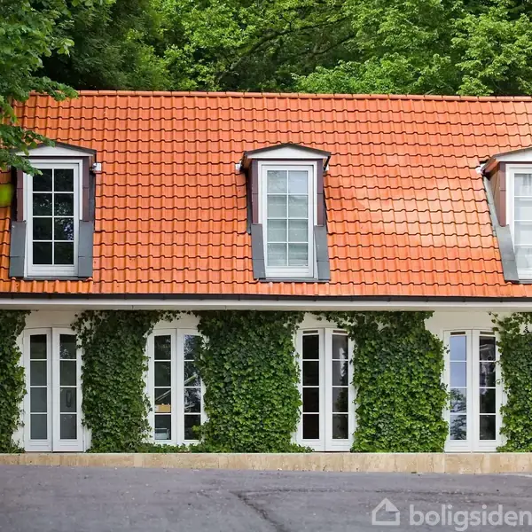 Hus med rødt tegltag og tre kviste; dækket af grønne planter. Store vinduer og hvid facade. Omgivet af frodig skov.
