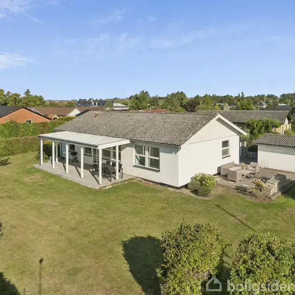 Huset står midt i en græsplæne, omkranset af hække under en blå himmel. Det har en overdækket terrasse og ligger i et beboelsesområde med andre huse i baggrunden.
