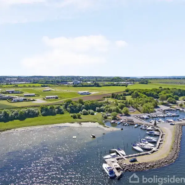 En kystnær marina med både fortøjet langs en mole. Bagved ses marker og spredte bygninger i et landligt område under en klar blå himmel.