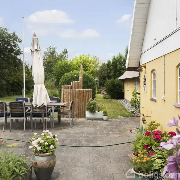 En terrasse med et bord og stole under to lukkede parasoller. Omkranset af planter og blomster i krukker. Gulmalet bygning til højre, grøn have og træer i baggrunden.