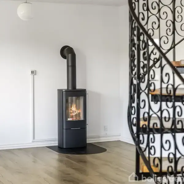 En moderne brændeovn står i hjørnet af et lyst rum med lyst trægulv. En elegant jerntrappe med dekorative mønstre ses i forgrunden.