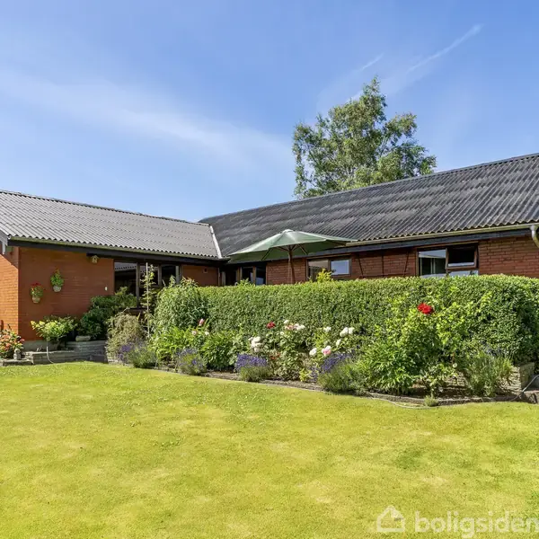 En rød murstensvilla med sort tag ligger omgivet af en velplejet have. Hække og blomsterbede skaber en farverig grænse, mens en parasol dækker en terrassesektion.