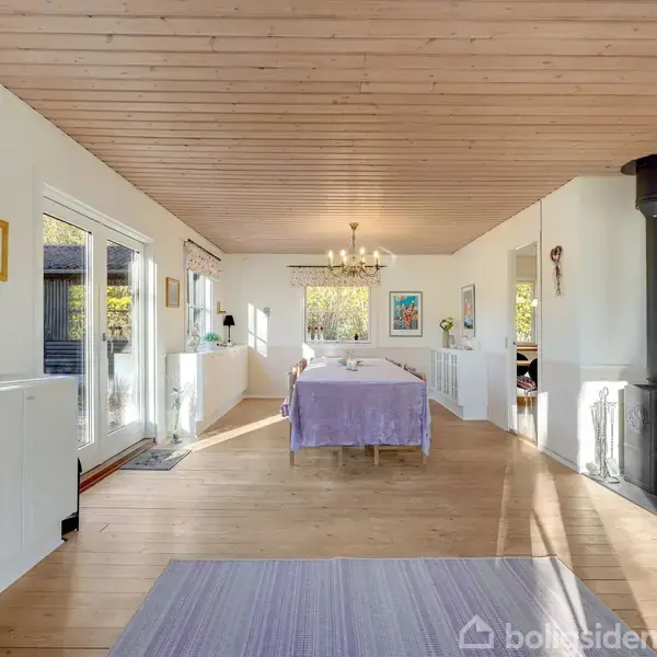 En brændeovn står ved væggen og opvarmer stuen med træloft og trægulv. Et spisebord med lilla dug står i midten, omgivet af store vinduer og dekorationer.