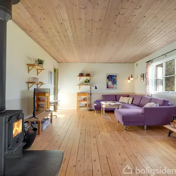 Brændeovn opvarmer stue med lilla hjørnesofa, trægulve og træloft. Blomster og billeder dekorerer rummet, der fyldes med naturligt lys fra vinduer med blomstrede gardiner.