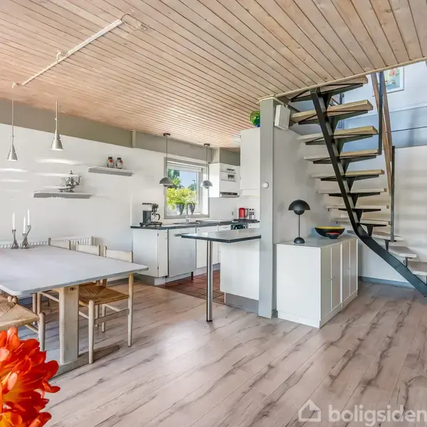 Spisestue med træbord og otte stole, åben køkkenområde med moderne apparater, trappe i mørkt træ fører opad. Rummets lyse trægulv og lofter skaber en indbydende atmosfære.