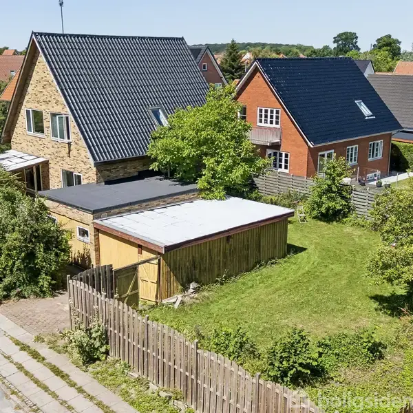 Et muret hus med sort tag står ved siden af en lille bygning i en forstadshave. Der er flere lignende huse i baggrunden og træer omkring plænen.