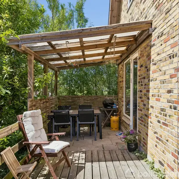 Træmøbler står på en overdækket terrasse ved siden af en murstensbygning. På terrassen er der også et havebord med stole. Omgivet af frodigt grønt løv og overskyet af en træpergola.