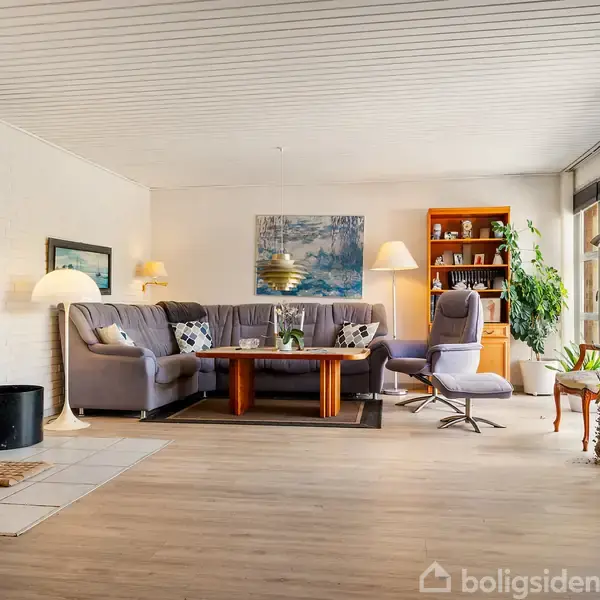 Stue med grå hjørnesofa og træbord, flankeret af en brændeovn til venstre. Hylder og planter bagved, store vinduer til højre slipper lys ind.