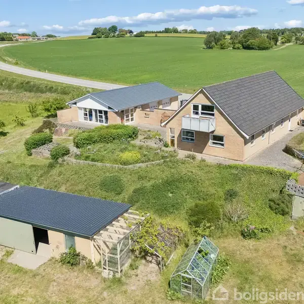 Et gult murstenshus med sort tag og store vinduer står i et åbent, grønt landskab. Et mindre skur med grønt tag ses tæt på huset, omgivet af græs og små planter.