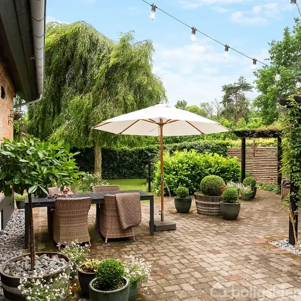 Terrasse med spisebord og stole under en parasol, omkranset af blomsterpotter og grønne buske, ved et murstenshus og sort skur, med dekorative lyskæder hængende over området.