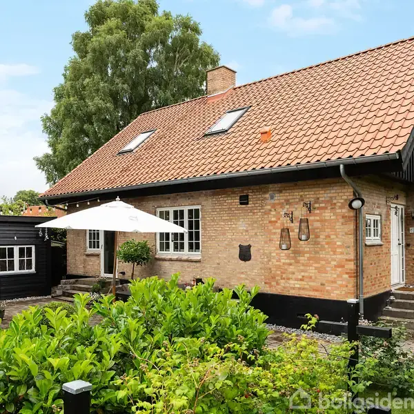 Et murstenshus med rødt tegltag har en terrasse med en hvid parasol. Omgivet af grønne buske og træer, står huset i en velplejet have.