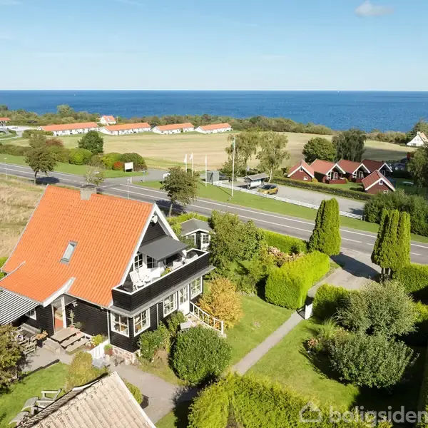 Et hus med rødt tag står i en velplejet have ved en landevej. Omkringliggende marker og huse strækker sig mod havet i baggrunden under en blå himmel.