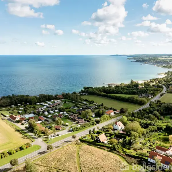 Kystby set fra oven med spredte huse og marker langs en snoet vej, der fører til en stor, blå havoverflade. Skovområder omkranser bebyggelsen under en blå himmel med skyer.