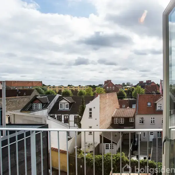 Åben altandør afslører byudsigt med række af traditionelle huse og grønne træer i baggrunden under delvist overskyet himmel.