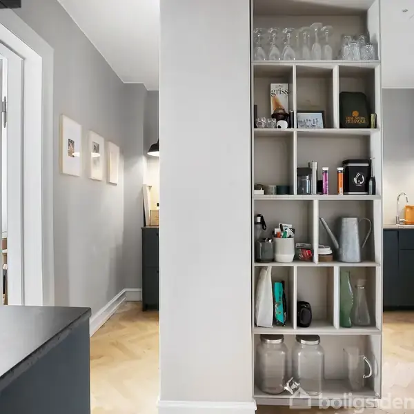 En reol fyldt med glas og bøtter står i en moderne, lys lejlighed. Til venstre ses en åben dør til soveværelset, og til højre er et køkkenområde synligt.