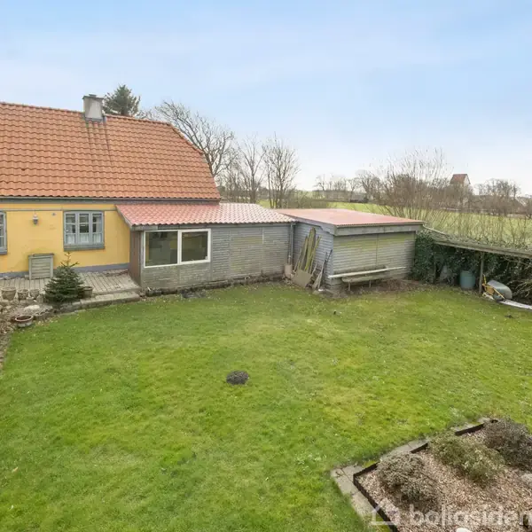Gult hus med rødt tegltag, stående i en grøn have med græsplæne og buske. Et skur står ved siden af huset. Træer og marker i baggrunden.