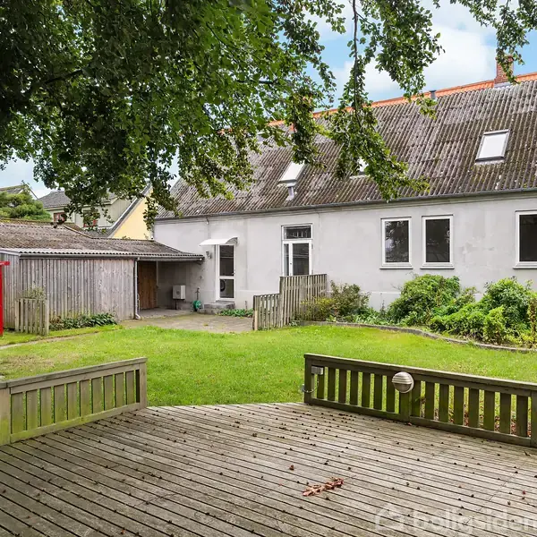 Træterrasse foran græsplæne og hvidt hus, omgivet af træer og planter. Huset har skråt tag med tagvinduer og er placeret i en rolig, grøn have.